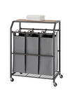 A laundry cart with three gray fabric bins hanging beneath a wooden top, all encased in a dark metal frame with wheels. A folded white towel rests on the wooden surface.