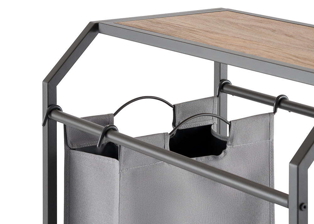 A close-up shot of a laundry cart with three gray fabric bags hanging from metal rods. The cart features a wooden top and a dark metal frame.
