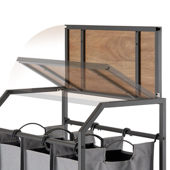 A close-up shot of a laundry cart with three gray fabric bags hanging below a metal frame. The cart features a flip-up wooden top, shown partially open with motion blur to indicate its hinged movement.
