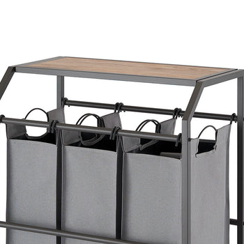 A close-up shot of a laundry cart with three gray fabric bags hanging from a metal frame. The cart features a wooden top surface and is set against a white background.