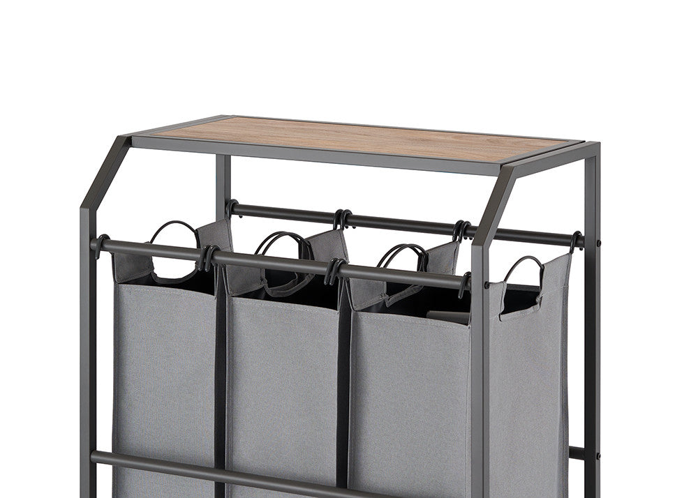 A close-up shot of a laundry cart with three gray fabric bags hanging from a metal frame. The cart features a wooden top surface and is set against a white background.