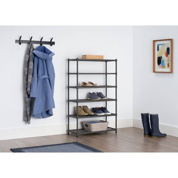 A six-tier metal slat shoe rack stands against a white wall, holding various pairs of shoes and a fabric storage bin on the bottom shelf. To the left, a black wall-mounted coat rack displays a blue coat and a gray scarf, while a framed abstract painting hangs on the right wall.