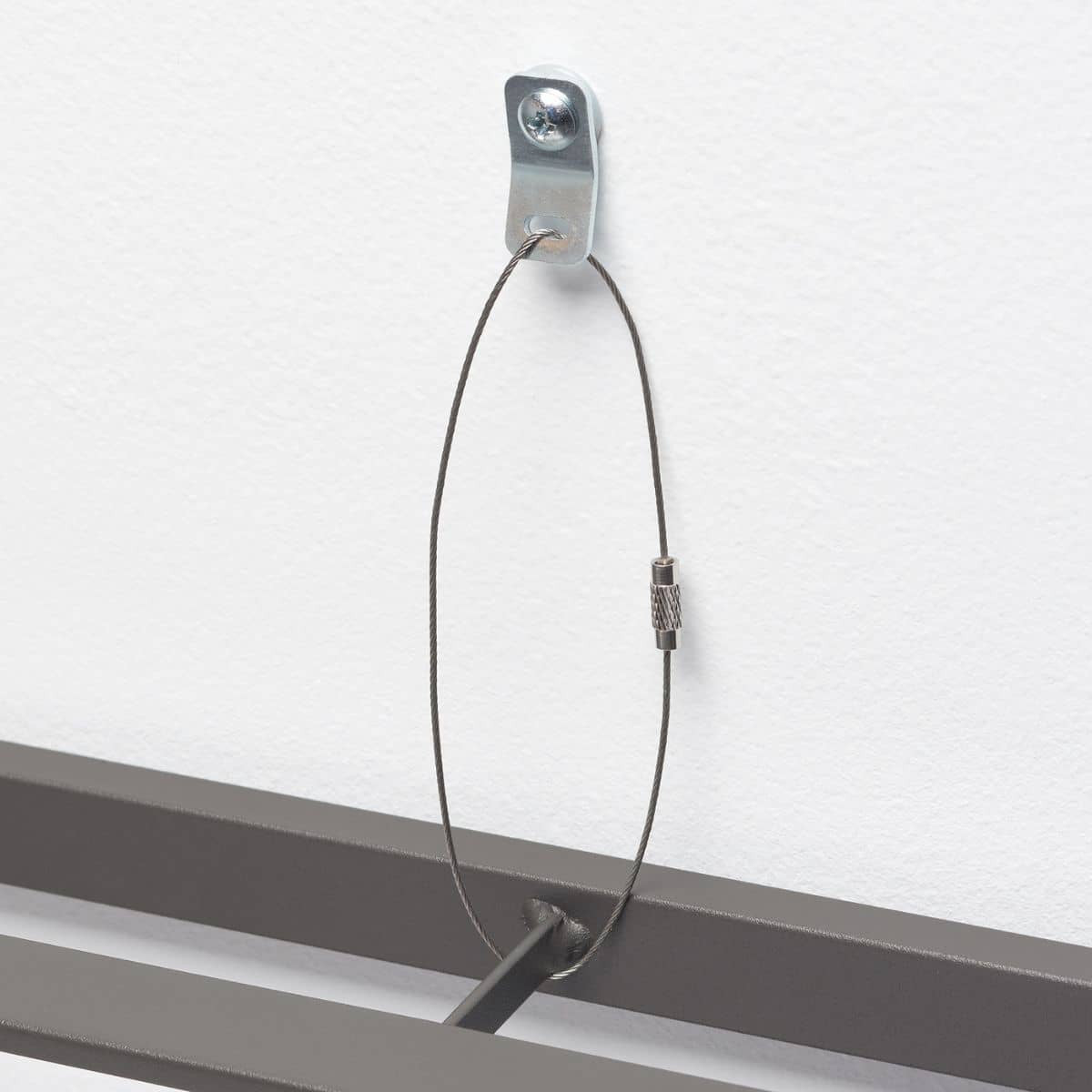 A close-up shows a thin, flexible steel cable forming a loop. One end of the cable is secured to a silver metal bracket screwed into a white wall, while the other end is attached to a dark gray metal frame.
