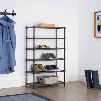 A black metal shoe rack with five shelves is neatly arranged in a modern entryway, holding various pairs of shoes and a fabric storage bin. A blue coat hangs on a wall-mounted hook, and a pair of navy rain boots stand on the wooden floor nearby.