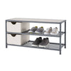 A three-tiered slate gray metal shoe bench with a light-colored top is shown against a white background. The middle and bottom shelves have horizontal slats and hold several pairs of shoes, while the top shelf also has two light-colored fabric storage baskets on the left side.