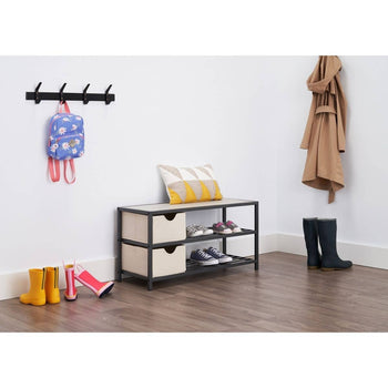 A mudroom or entryway is shown with a three-tiered shoe bench that has a light-colored wooden top, two light beige fabric drawers on the middle shelf, and a gray wire bottom shelf holding several pairs of shoes. Above the bench is a patterned pillow, and to the left, a black coat rack with a floral backpack hangs on it, while colorful rain boots sit on the floor below.