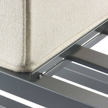 A close-up shot shows the corner of a light beige upholstered bed frame resting on a gray metal bed base with horizontal slats. The focus is on the textural contrast between the soft fabric and the rigid metal.