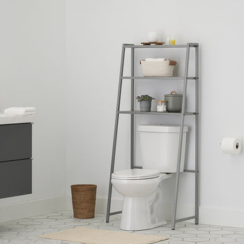 A metal over-the-toilet étagère with wooden shelves stands above a white toilet, filled with towels, toiletries, and a potted plant in a modern bathroom. The room features a dark gray wall-mounted vanity, a round gold-framed mirror, and grey hexagonal floor tiles.