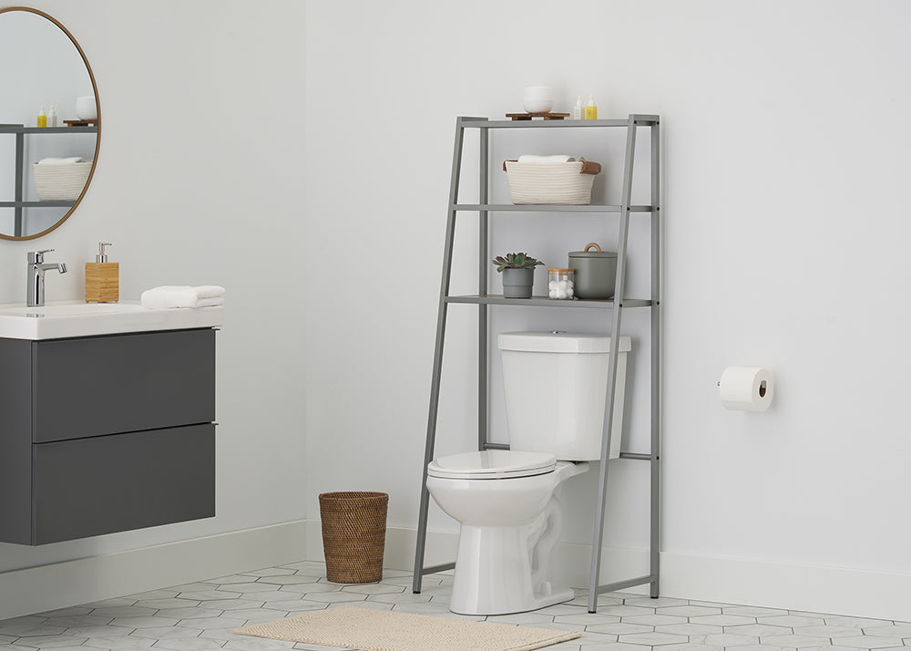 A metal over-the-toilet étagère with wooden shelves stands above a white toilet, filled with towels, toiletries, and a potted plant in a modern bathroom. The room features a dark gray wall-mounted vanity, a round gold-framed mirror, and grey hexagonal floor tiles.