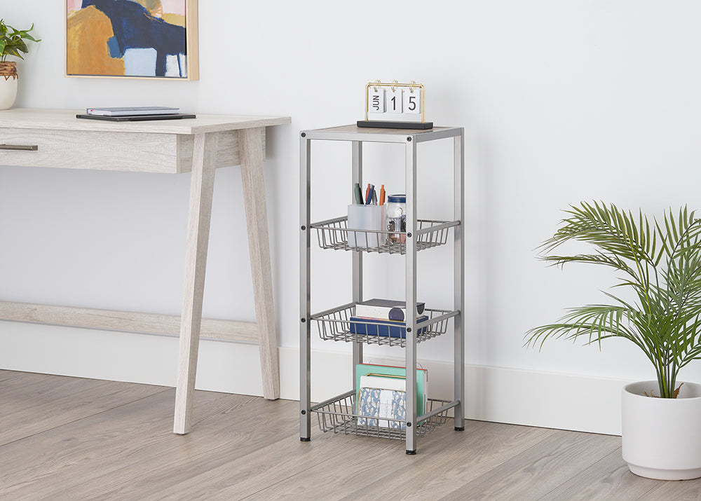 A light-colored, tall, rectangular shelf unit with three wire baskets is positioned next to a matching desk. The top shelf holds pens and a calendar, while the baskets contain various office supplies and documents.