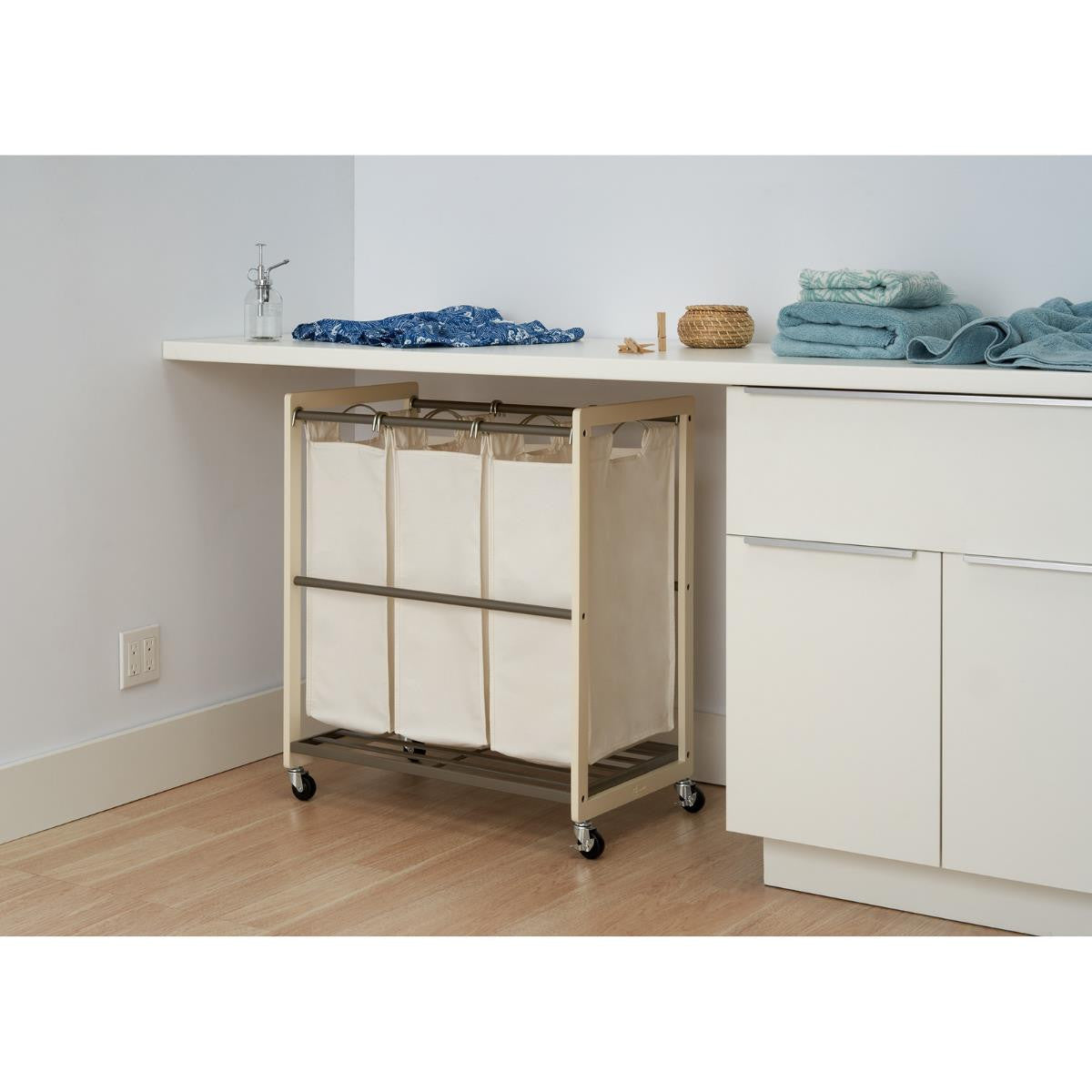 A white Trinity 3-Bag Laundry Cart with a tabletop is positioned beneath a white counter in a laundry room. The cart features three removable canvas bags and wheels for easy mobility.
