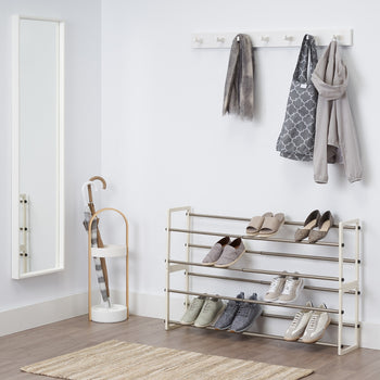 A well-lit entryway features a four-tiered metal and white-colored wood shoe rack holding various pairs of shoes. Above the rack, a white wall-mounted coat rack displays a gray scarf, a patterned tote bag, and a light jacket. To the left, a tall mirror leans against the wall next to a white umbrella stand with two umbrellas. A woven rug lies on the wooden floor in front of the shoe rack.