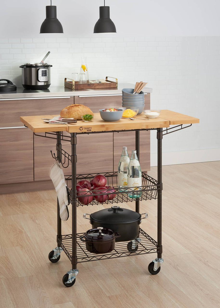 A bronze metal kitchen cart with a light wood butcher block top stands in a kitchen. The cart has two wire shelves holding produce, water bottles, and cookware, and hanging hooks with an oven mitt.