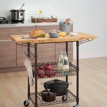 A bronze metal kitchen cart with a light wood butcher block top stands in a kitchen. The cart has two wire shelves holding produce, water bottles, and cookware, and hanging hooks with an oven mitt.