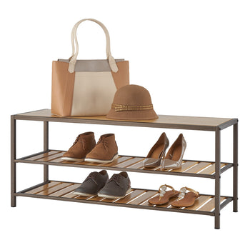 A three-tiered bamboo shoe organizer with a bronze metal frame holds various footwear and accessories. On the top shelf, a tan and beige tote bag and a brown hat with gold embellishments sit next to two pairs of shoes on the middle shelf: brown suede ankle boots and glossy brown heels. The bottom shelf contains two pairs of shoes: dark brown casual shoes and brown sandals with decorative stones.