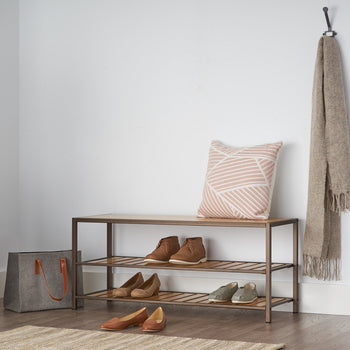 A three-tiered bronze anthracite shoe rack with a wooden bench top sits against a white wall. Shoes are neatly arranged on the rack, and a patterned pillow and a scarf add decorative touches.