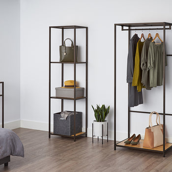 A tall, four-tiered metal shelf with bamboo surfaces stands against a white wall, holding a green purse, a gray box with a yellow hat, and a gray fabric bin. This shelving unit appears to be the Trinity 4-Tier Bamboo Shelving Tower, offering stylish and functional storage.