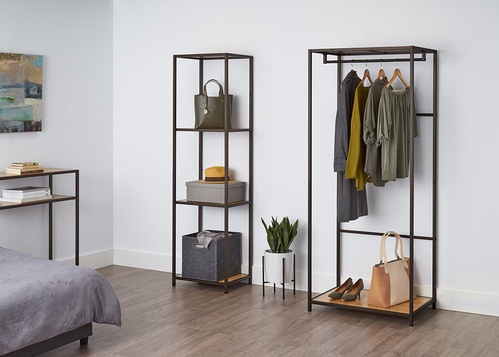 A tall, four-tiered metal shelf with bamboo surfaces stands against a white wall, holding a green purse, a gray box with a yellow hat, and a gray fabric bin. This shelving unit appears to be the Trinity 4-Tier Bamboo Shelving Tower, offering stylish and functional storage.