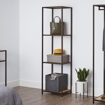 A tall, four-tiered metal shelf with bamboo surfaces stands against a white wall, holding a green purse, a gray box with a yellow hat, and a gray fabric bin. This shelving unit appears to be the Trinity 4-Tier Bamboo Shelving Tower, offering stylish and functional storage.