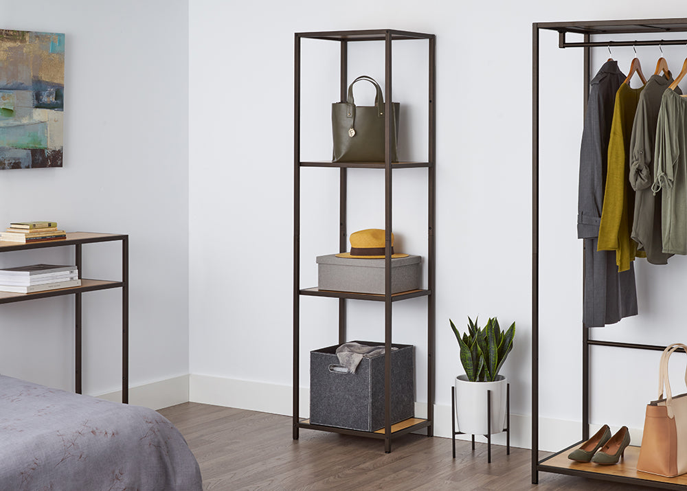 A tall, four-tiered metal shelf with bamboo surfaces stands against a white wall, holding a green purse, a gray box with a yellow hat, and a gray fabric bin. This shelving unit appears to be the Trinity 4-Tier Bamboo Shelving Tower, offering stylish and functional storage.