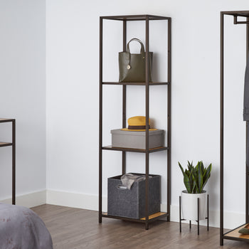 A tall, four-tiered metal shelf with bamboo surfaces stands against a white wall, holding a green purse, a gray box with a yellow hat, and a gray fabric bin. This shelving unit appears to be the Trinity 4-Tier Bamboo Shelving Tower, offering stylish and functional storage.