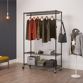 A Trinity Bronze Rolling Garment Rack, a closet organizer on wheels, holding several pieces of clothing. The rack has three wire shelves and a hanging rod, with garments hung and folded neatly on it, along with a handbag hanging from the side.