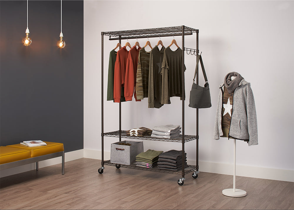 A Trinity Bronze Rolling Garment Rack, a closet organizer on wheels, holding several pieces of clothing. The rack has three wire shelves and a hanging rod, with garments hung and folded neatly on it, along with a handbag hanging from the side.