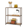 A three-tiered bronze anthracite shelving rack filled with kitchen items. The top shelf holds a white ceramic utensil holder with wooden kitchen tools and a black wire basket filled with yellow lemons. The middle shelf contains a wooden tray with a clear pitcher and two clear glasses. The bottom shelf holds a dark wire basket filled with books.