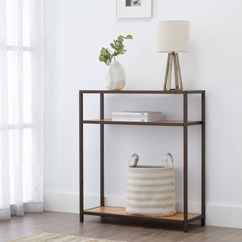 A bronze anthracite three-tiered rack used a a accent shelving unit in a living room. It has a lamp, a vase with greenery, and a striped basket adorn the table, complemented by a framed artwork on the wall.