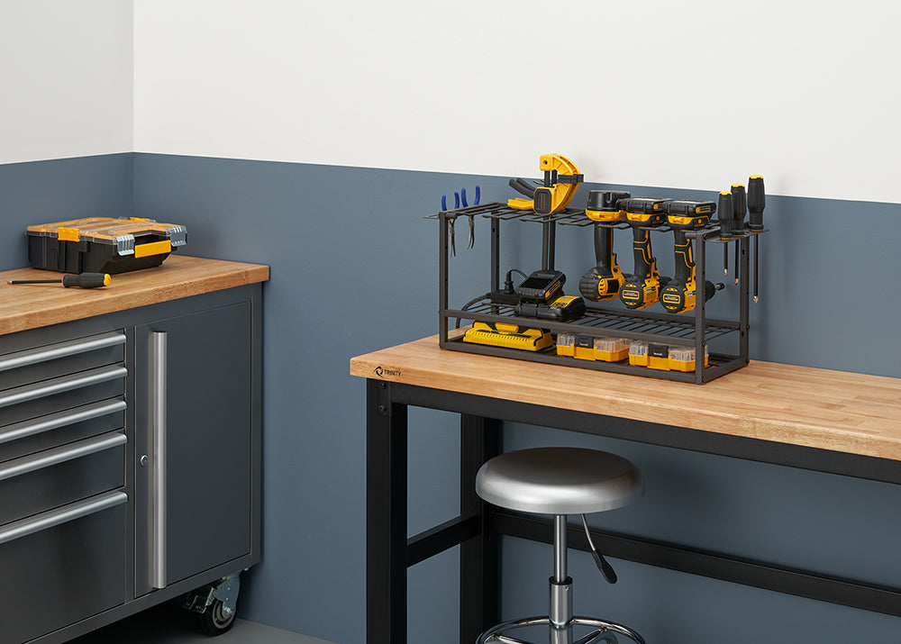 
A dark gray metal wire rack on a wooden workbench holds a collection of yellow and black power tools and accessories, including a sander and several drills. On the left, a matching rolling tool cabinet with a wooden top sits next to the workbench.