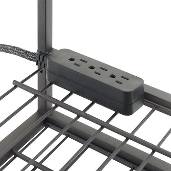 A black, rectangular power strip with three outlets is attached to the side of a metal wire shelf. The power strip's braided cord extends from the left, secured to the vertical metal frame with a tie.