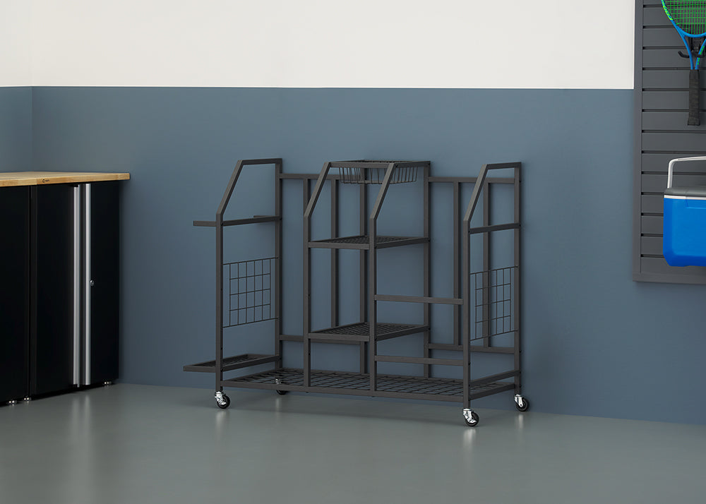 A black metal rolling storage cart stands on a garage floor against a blue and white wall. The multi-tiered organizer features various shelves and slots, next to a black cabinet with a wood countertop.