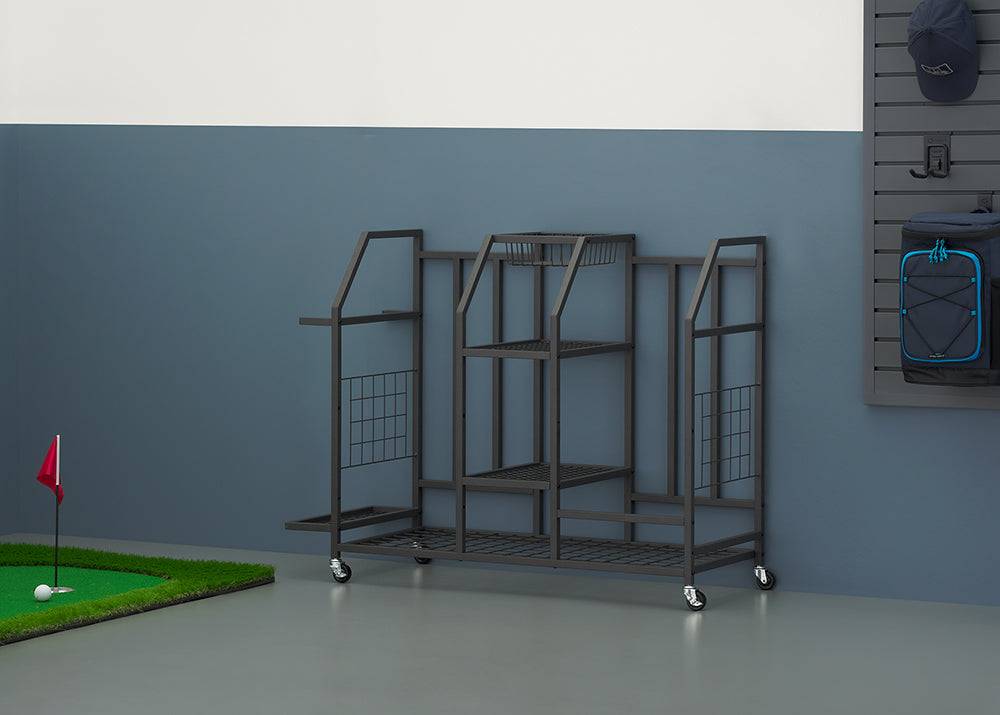 A black metal rolling storage rack with multiple tiered shelves and partitions stands on a garage floor against a blue-grey wall. The multi-purpose organizer is next to a indoor putting green with a ball and flag on the left, and a blue backpack hangs from a slat wall panel to the right.