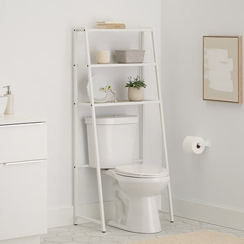 A white metal over-the-toilet space saver with wood slatted shelves is positioned over a toilet in a light-filled bathroom. The space saver shelves are styled with stacked towels, a small decorative box, a ceramic container, a framed mirror, and a small potted plant.