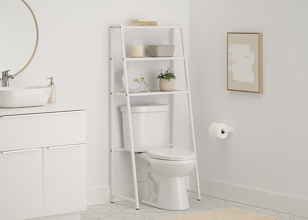 A white metal over-the-toilet space saver with wood slatted shelves is positioned over a toilet in a light-filled bathroom. The space saver shelves are styled with stacked towels, a small decorative box, a ceramic container, a framed mirror, and a small potted plant.