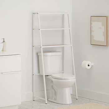 A leaning white metal A-frame over-the-toilet space saver stands above a white toilet in a clean, modern bathroom with grey hexagon tile flooring. The space saver features two slatted wood-toned shelves which are empty, and the surrounding scene includes a white vanity, a round mirror, and a small abstract painting.
