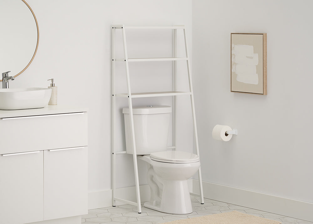 A leaning white metal A-frame over-the-toilet space saver stands above a white toilet in a clean, modern bathroom with grey hexagon tile flooring. The space saver features two slatted wood-toned shelves which are empty, and the surrounding scene includes a white vanity, a round mirror, and a small abstract painting.