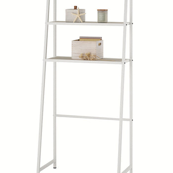 A white metal, leaning over-the-toilet étagère is displayed against a plain white background, featuring three shelves with light-wood slats. The middle shelf holds a wooden storage box with files, while the top shelf displays a starfish, a candle, and other small decor items.