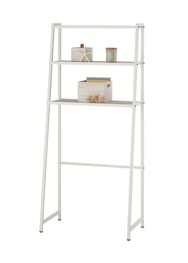 A white metal, leaning over-the-toilet étagère is displayed against a plain white background, featuring three shelves with light-wood slats. The middle shelf holds a wooden storage box with files, while the top shelf displays a starfish, a candle, and other small decor items.