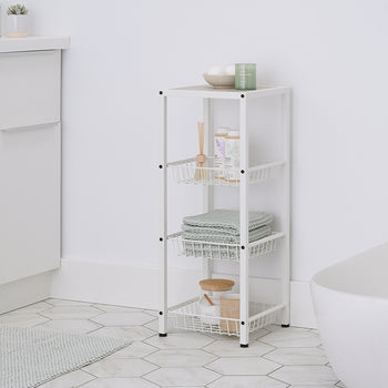 A four-tiered, white metal storage cart with pull-out wire baskets stands in a modern bathroom. The top shelf holds bath bombs and a candle, while the baskets below contain folded green towels, toiletries, and a wooden jar with bath salts.