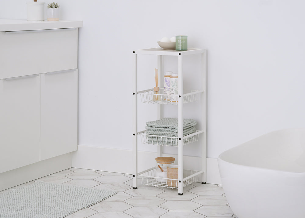 A four-tiered, white metal storage cart with pull-out wire baskets stands in a modern bathroom. The top shelf holds bath bombs and a candle, while the baskets below contain folded green towels, toiletries, and a wooden jar with bath salts.