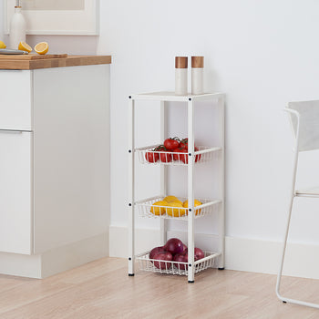A narrow, white four-tiered metal cart with mesh baskets holds red tomatoes, yellow lemons, and red onions. The cart stands on a light wood floor in a white kitchen, next to a white cabinet with a wooden countertop.
