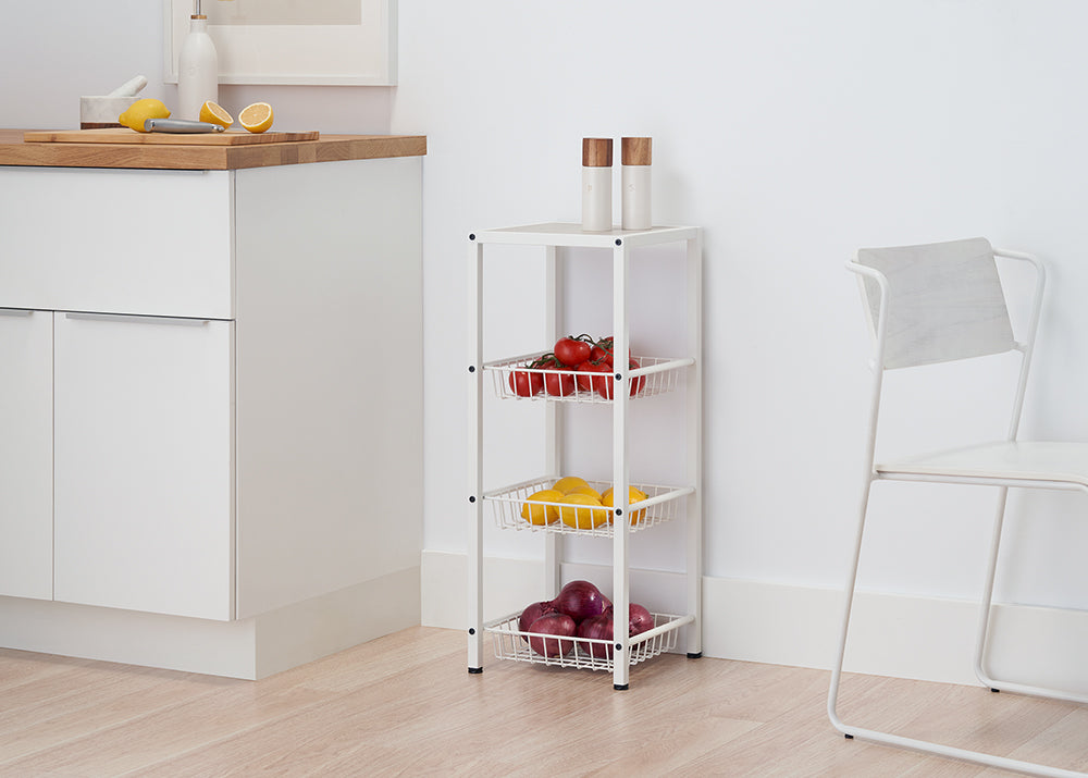A narrow, white four-tiered metal cart with mesh baskets holds red tomatoes, yellow lemons, and red onions. The cart stands on a light wood floor in a white kitchen, next to a white cabinet with a wooden countertop.