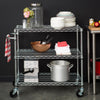A three-tiered chrome wire rolling kitchen cart holds various kitchen and dining items against a dark background. The cart contains dishes, bowls, a copper mixing bowl with utensils, towels, a small espresso machine, and a large stockpot.