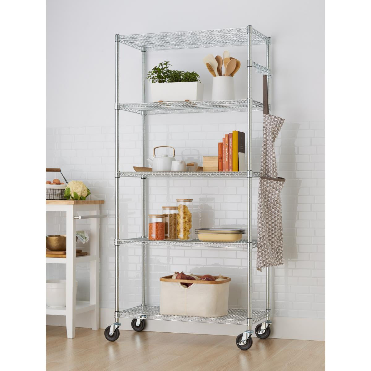 A tall, five-tier chrome wire shelving rack with wheels stands against a white subway tile wall. The shelves hold various kitchen and household items, including plants, cookbooks, pantry staples in jars, and a cloth hanging from the side.