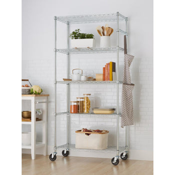A tall, five-tier chrome wire shelving rack with wheels stands against a white subway tile wall. The shelves hold various kitchen and household items, including plants, cookbooks, pantry staples in jars, and a cloth hanging from the side.