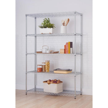 A tall, five-tier silver metal wire shelving unit stands against a white tiled wall in a kitchen. The shelves hold various kitchen items including plants, utensils, teaware, books, jars of food, a baking pan, and a cloth bin filled with produce.
