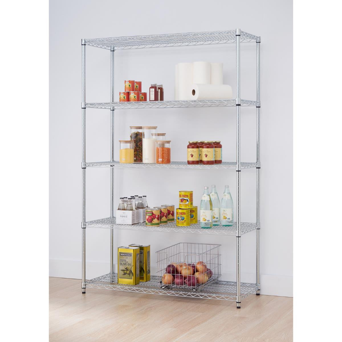 A tall, five-tier wire shelving rack stands on a kitchen with a light-colored wooden floor. The shelves hold various pantry items, including canned goods, jars of food, paper towels, bottles, and a wire basket filled with red onions.