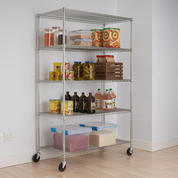 A tall, five-tier chrome wire shelving rack on wheels is shown against a white wall and light-colored floor. The shelves hold various pantry items including jars of lentils and spices, cereal boxes, canned goods, bottles of oil, and large clear containers of dried beans and rice.