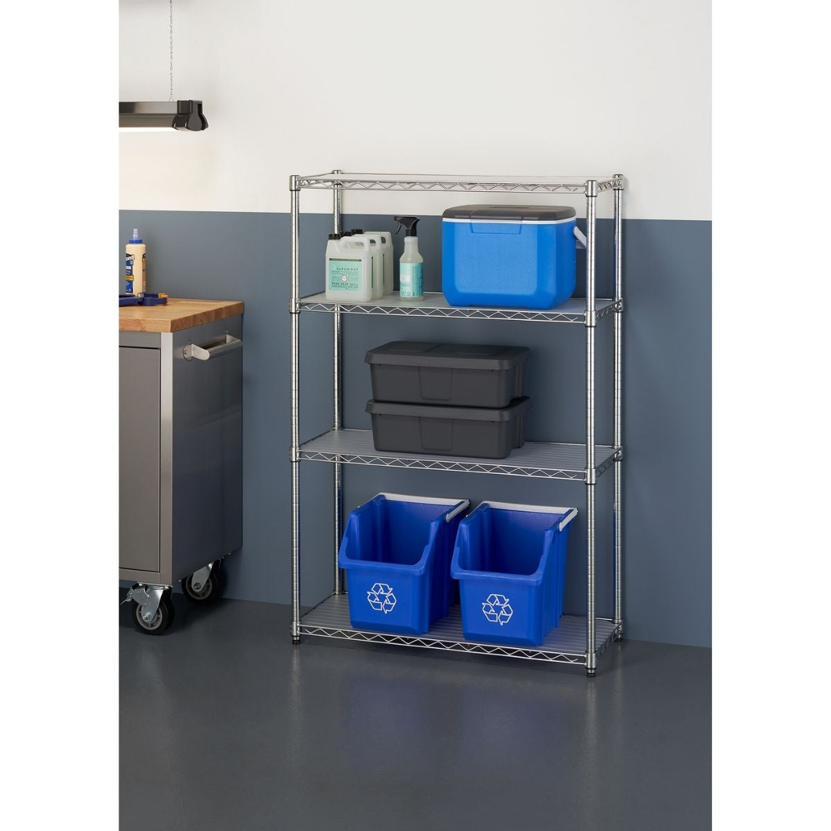 A four-tier wire shelving rack stands against a two-toned wall, holding various storage items. The shelves contain a blue cooler, cleaning supplies, two dark gray storage containers, and two blue recycling bins.